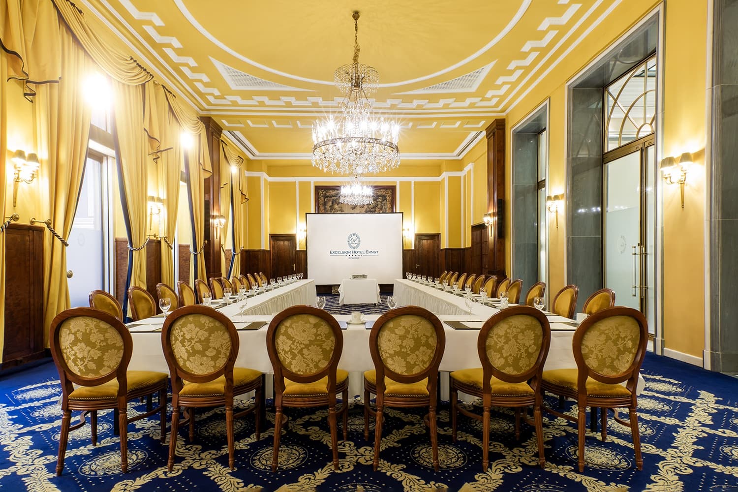 In einem Tagungsraum im Tagungshotel Köln mit blauem Teppich und gelben Vorhängen steht eine Tafel in U-Form