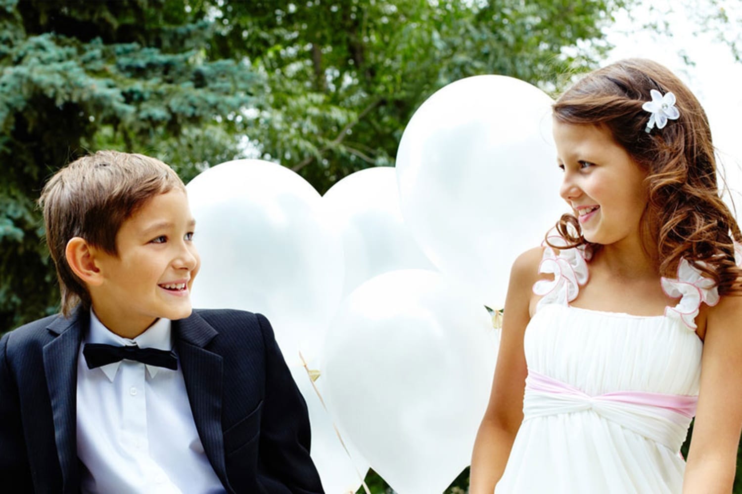 Zwei Kinder tragen festliche Kleidung und stehen draußen vor weißen Luftballons
