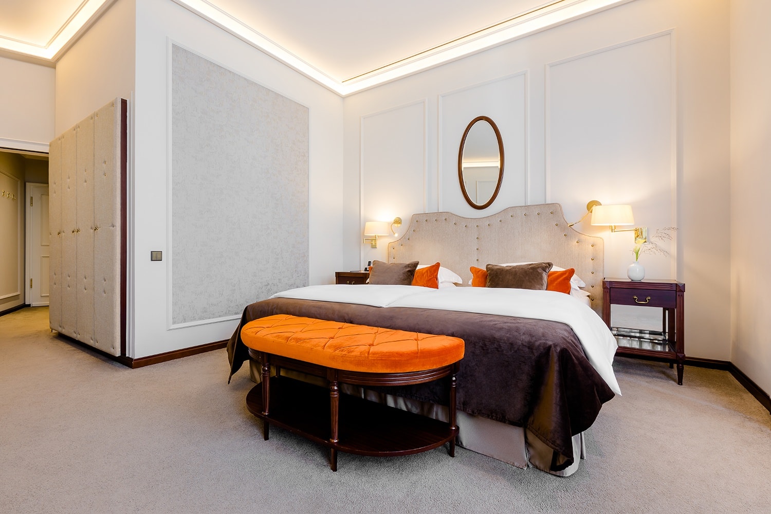 Großes Doppelbett steht auf einem Teppichboden vor einem ovalen Spiegel und zwischen zwei Nachttischen